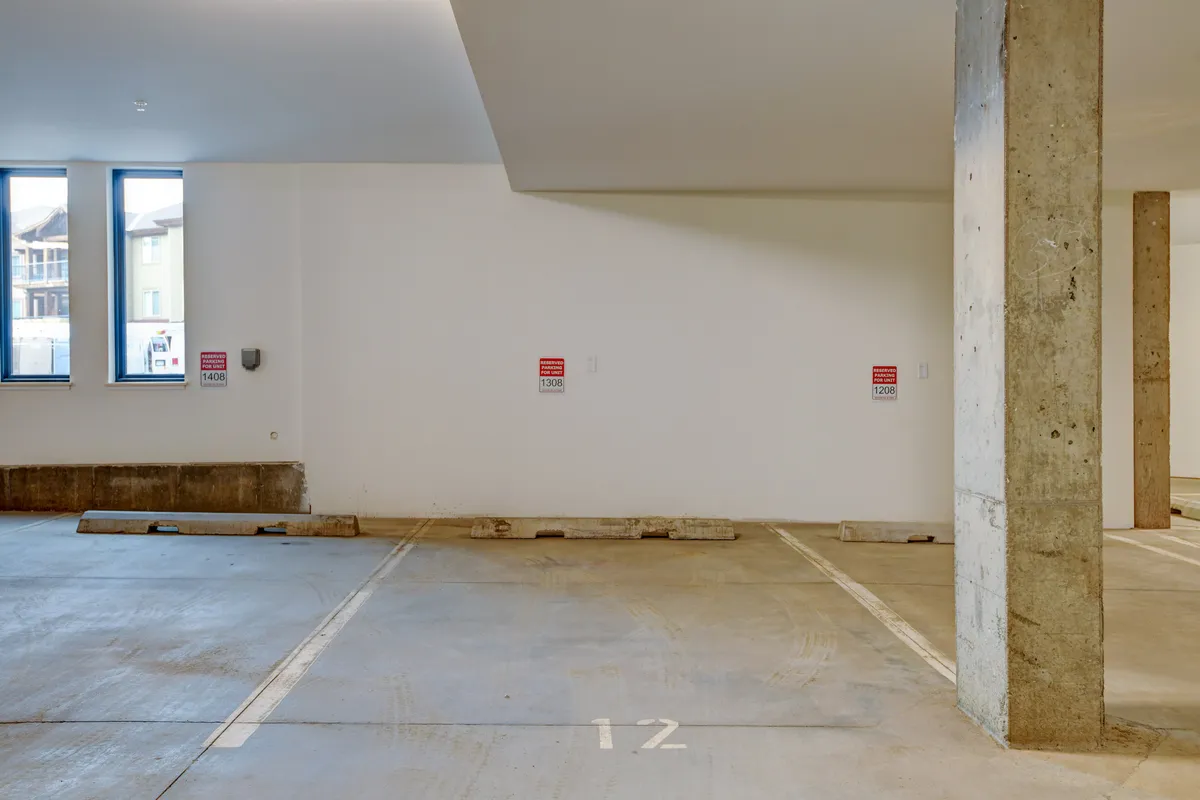 An interior view of a well-lit parking garage with empty spaces, concrete pillars, and natural light streaming through windows. The area appears spacious and safe, inviting for parking vehicles.