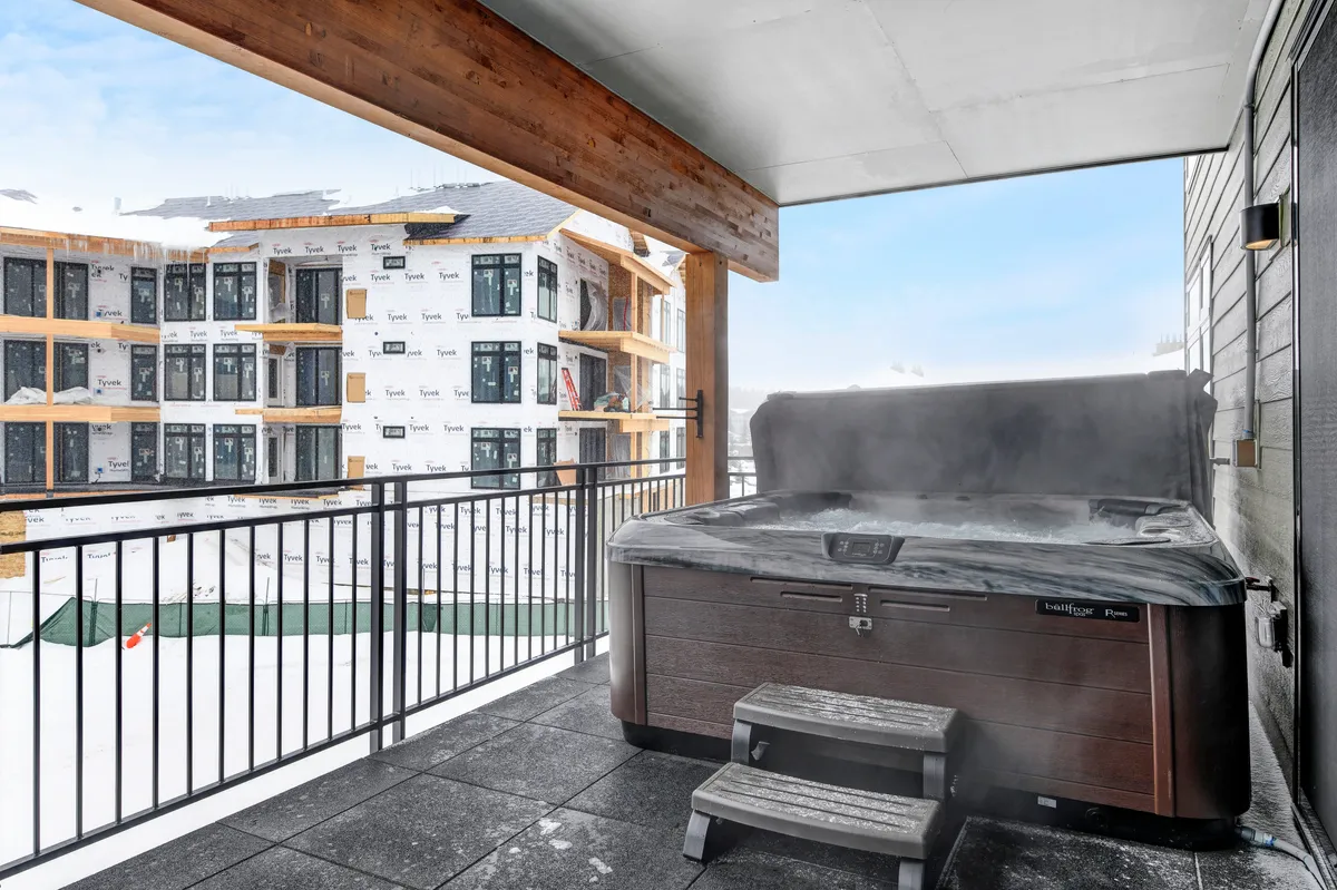 An exterior shot of a private hot tub set against a snowy landscape, with steam rising and snow covering the surroundings. The hot tub is located on a balcony or patio, offering a cozy retreat with views of the wintry outdoors, and subtle hints of ongoing development in the area.