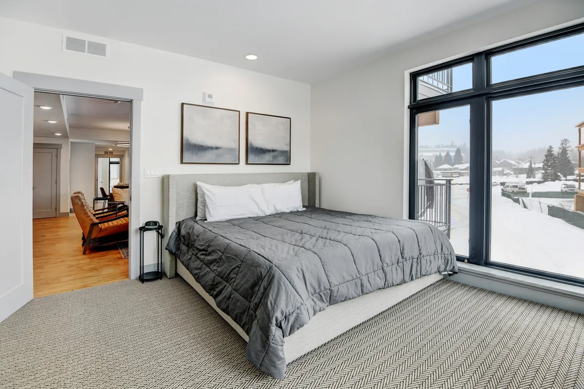 This image features a bed with gray bedding positioned near large windows, allowing abundant natural light to filter into the room. Wall art adds a touch of personality, and the tranquil setting evokes a sense of restful escape.