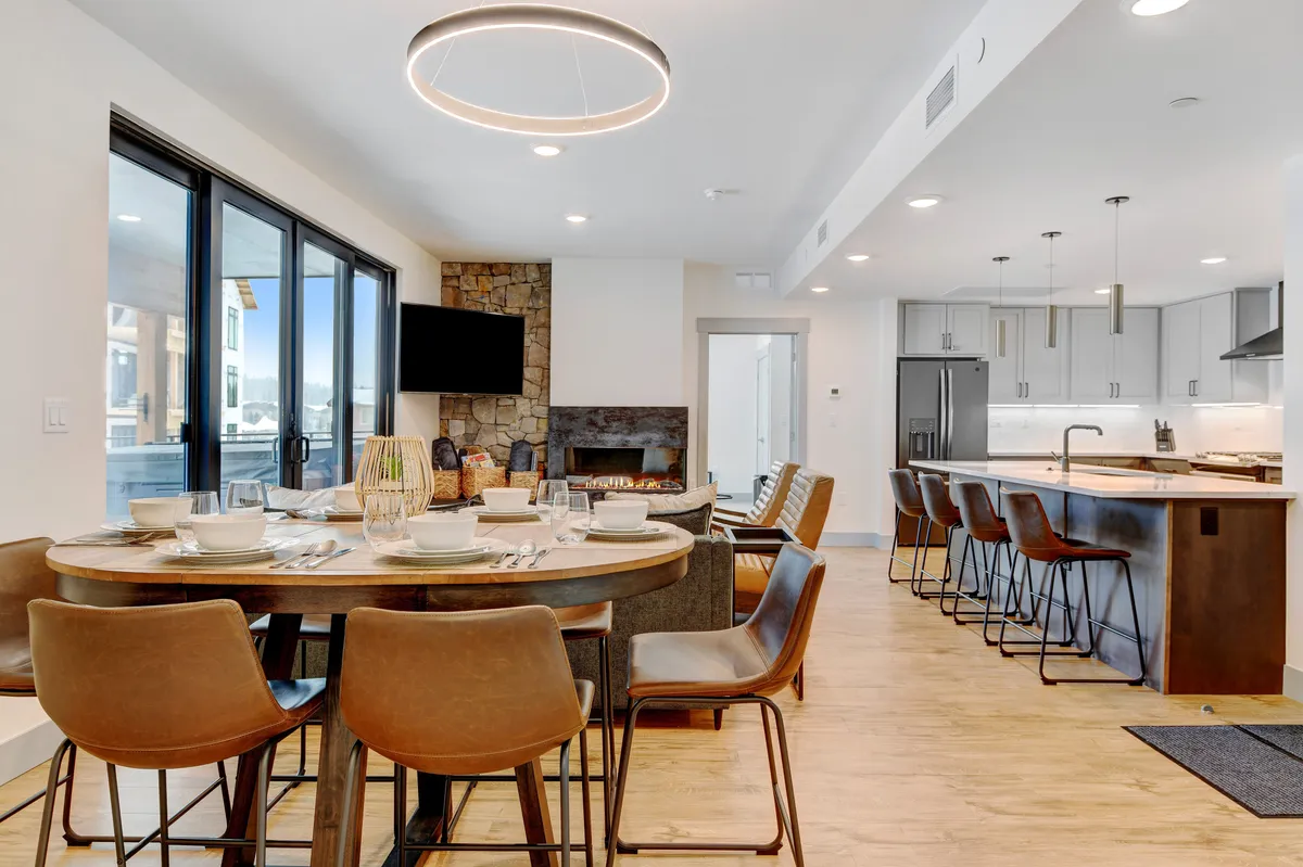 A stylish dining area set for a meal, adjacent to a modern kitchen with a prominent granite island. Large windows fill the room with natural light, and a stone-surround fireplace anchors the open-concept layout, creating an inviting space for gatherings.