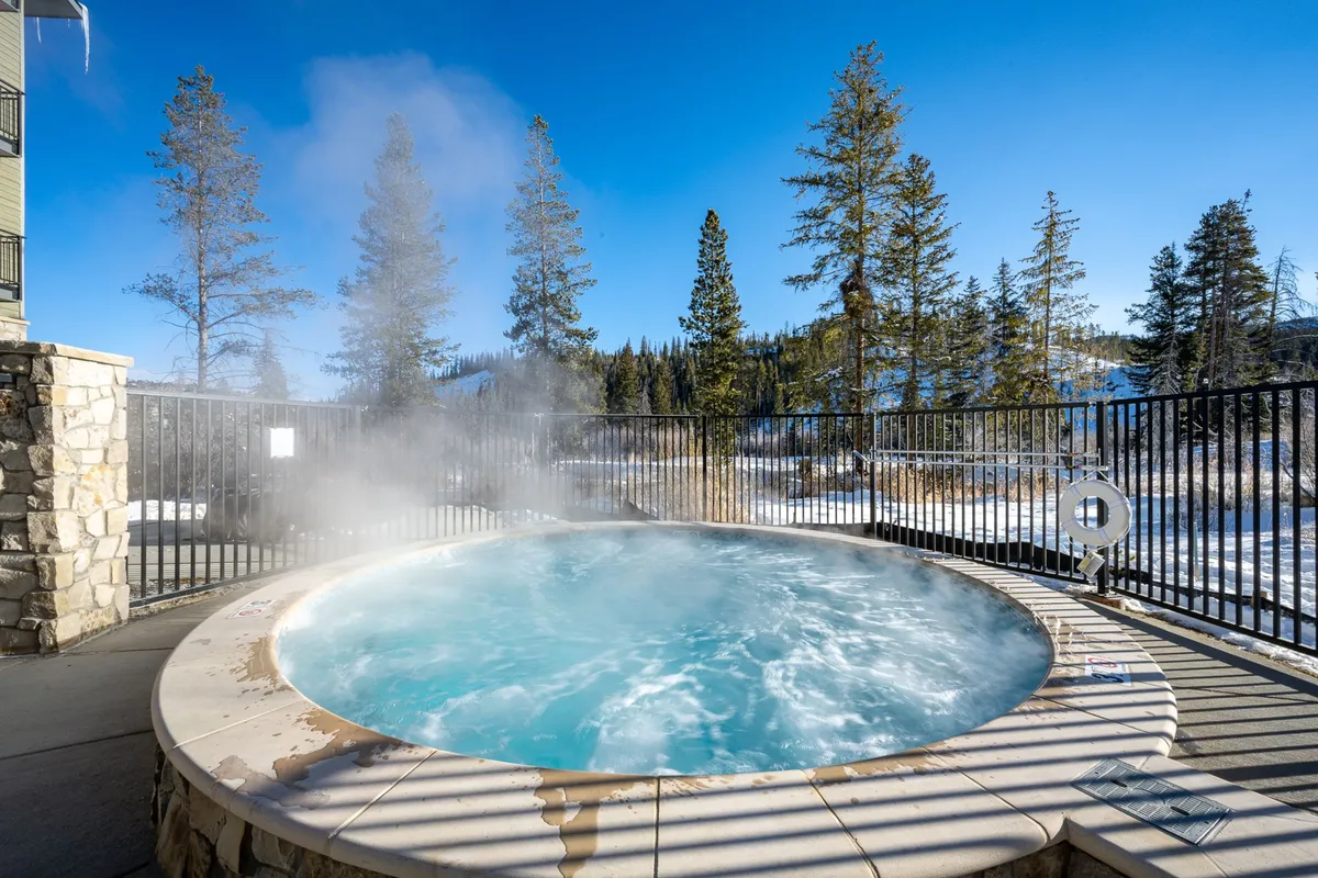A serene outdoor hot tub surrounded by lush trees, with clear blue skies overhead. The natural setting is inviting and tranquil, suggesting a private, relaxing soak while being immersed in the beauty of the mountains.