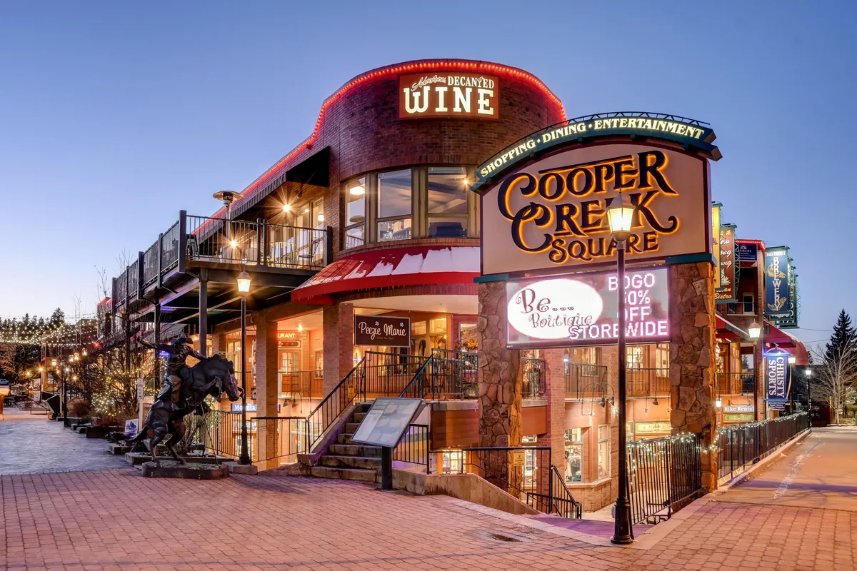 Cooper Creek Square, marked by its prominent sign, wine shop, staircase leading to the upper level, outdoor seating, and a distinctive horse sculpture. The scene is bustling with possibilities for dining, shopping, and socializing, reflecting the heart of downtown Winter Park.