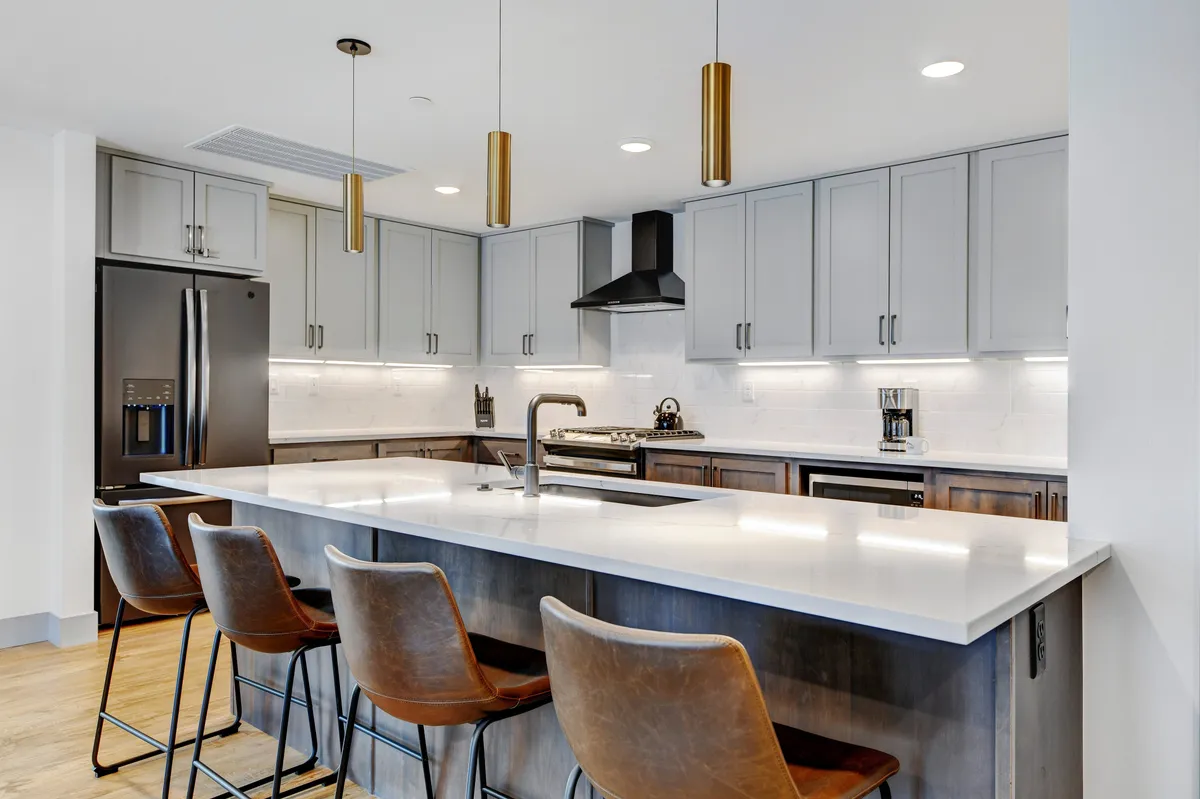 The image showcases a sleek, modern kitchen with a spacious island surrounded by stylish bar stools, gleaming stainless steel appliances, and contemporary lighting fixtures. The open, airy layout hints at a welcoming space perfect for gathering, cooking, and enjoying time together.