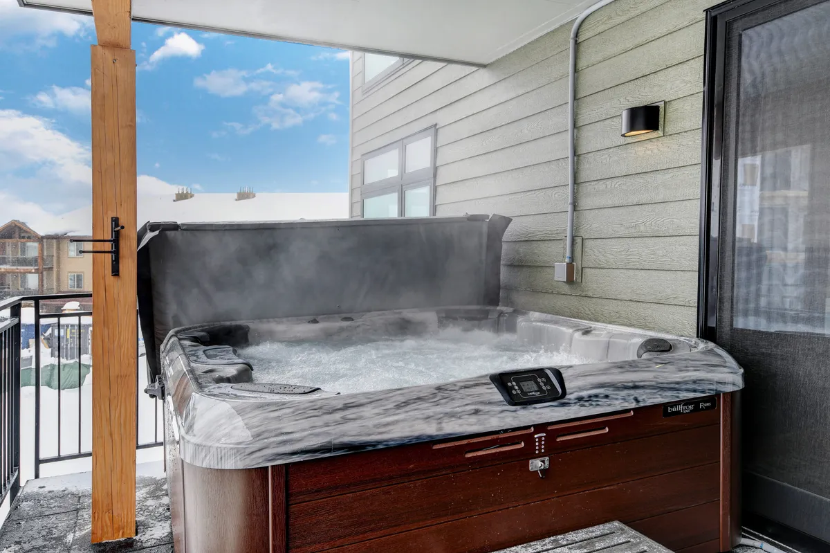 A private outdoor patio featuring a bubbling hot tub surrounded by a snowy mountain landscape and a wooden structure. Guests are relaxing in the hot tub, enjoying the peaceful views and serene mountain vibes after a day of skiing or hiking.