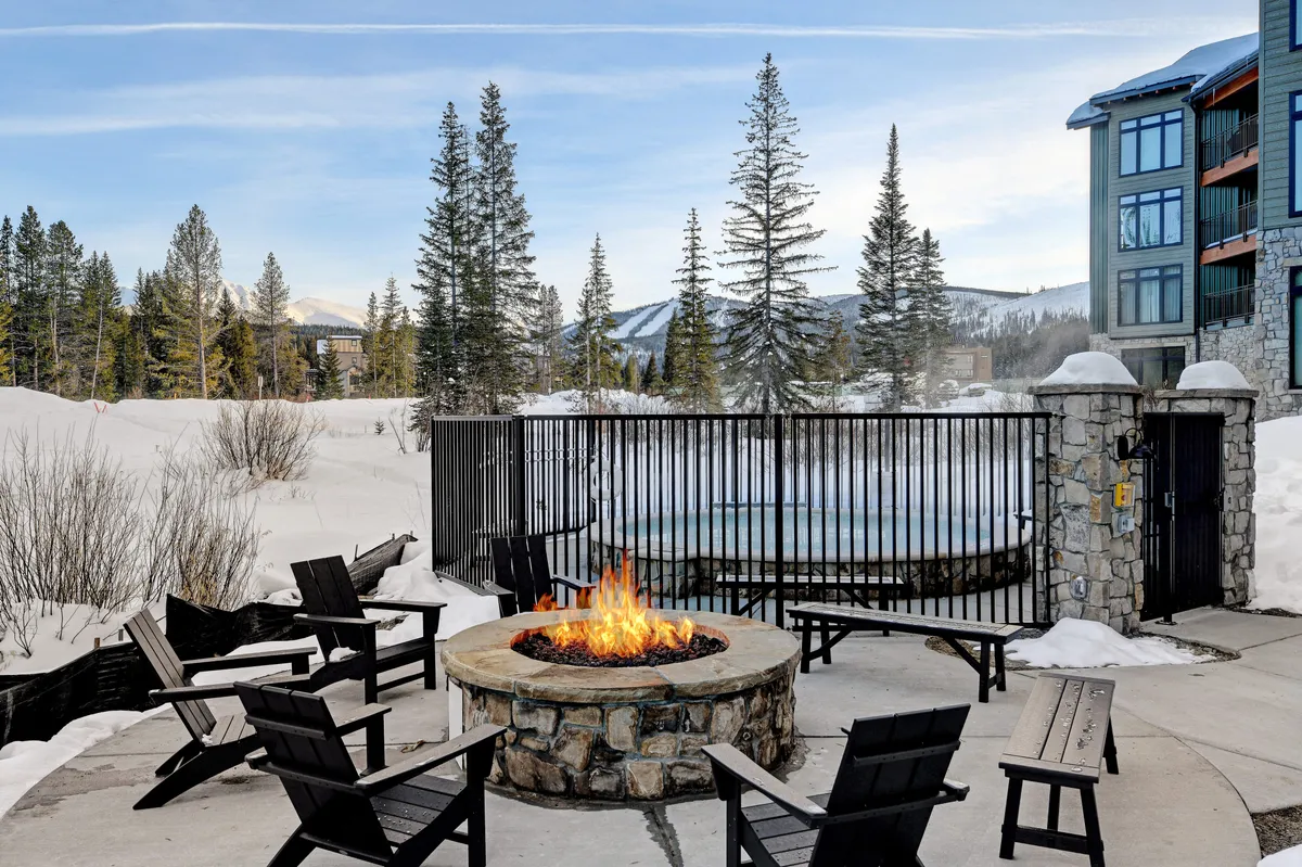 A cozy outdoor community firepit area with surrounding chairs, set against a snow-covered landscape and distant mountain peaks. Friends are gathered around the fire, sipping hot beverages and enjoying the warmth after a day outdoors.