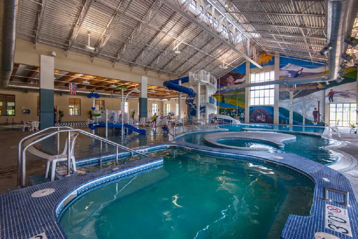 A vibrant indoor recreation center with a swimming pool, water slide, and children playing beneath a colorful mural. The atmosphere is lively and joyful, offering plenty of energy for family fun and rejuvenation.