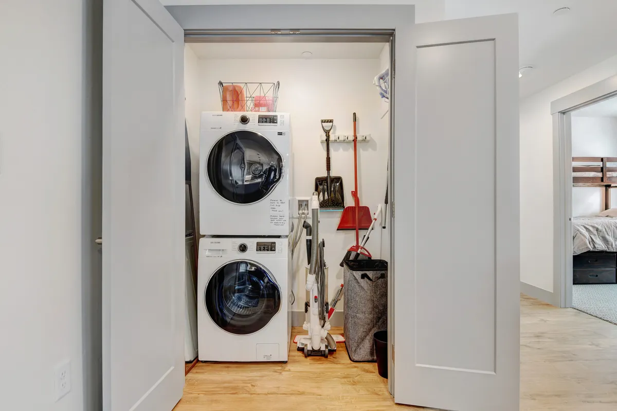 A compact laundry area with a stacked washer and dryer, cleaning tools, and a storage basket. The practical space is organized and functional, ready for refreshing gear after a day outside.
