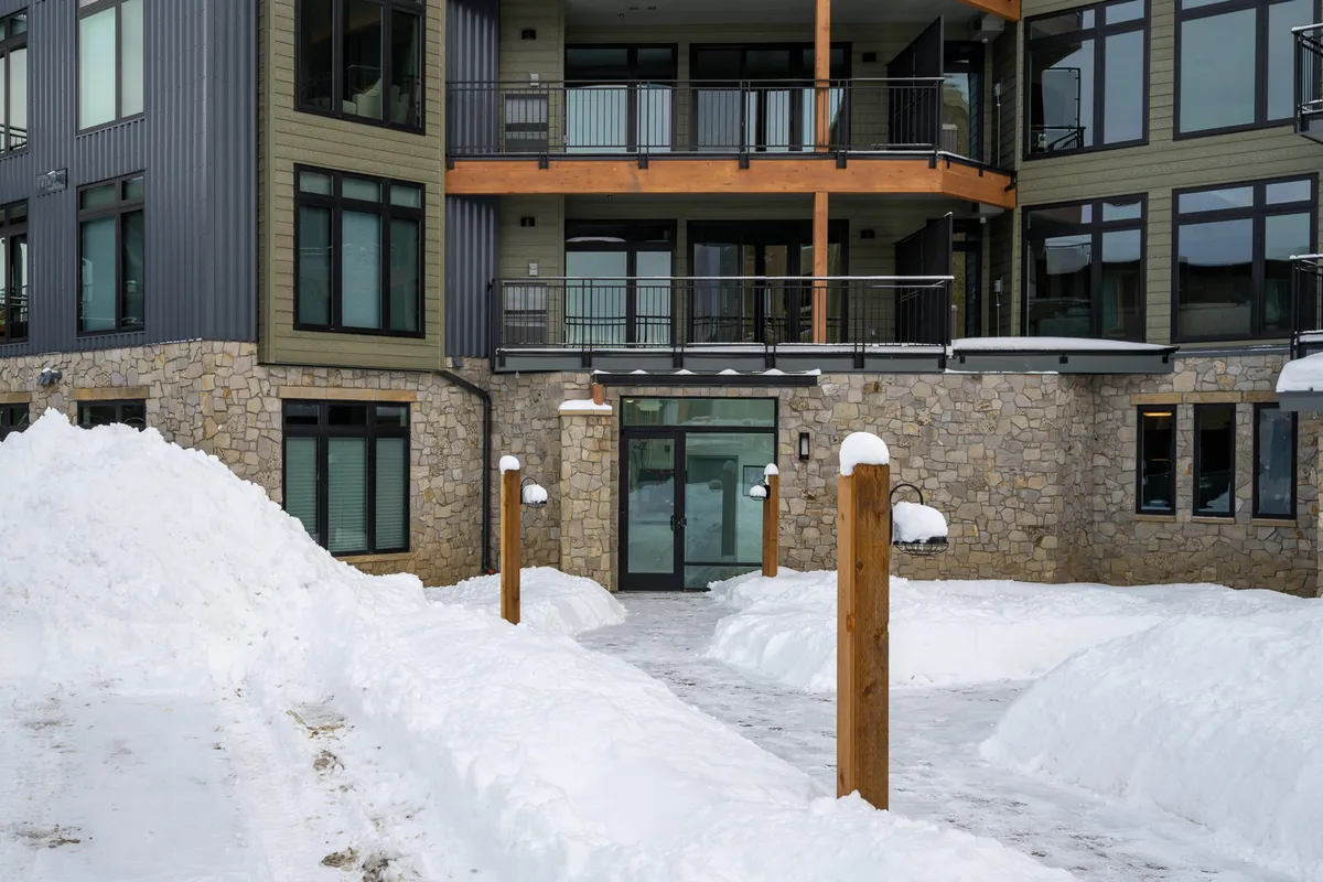 A serene exterior view of the condo, featuring snow-covered pathways, stone and wood architectural elements, and large windows. The setting evokes a peaceful neighborhood vibe and hints at the possibility of outdoor fun, such as building snowmen or taking family walks.